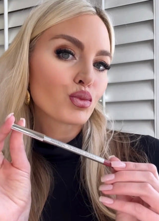 Model with long blonde hair wearing glam makeup, holding Mirabella’s Lip Definer pencil in shade Sassy and showcasing a defined nude-rose lip look.