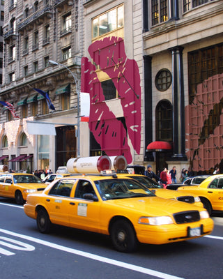 Yellow taxis with oversized Mirabella lipstick ads drive through a city street, with bold lipstick wall art creating a fashion-forward urban vibe.