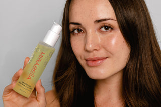 Model holding Mirabella Skinphony Pro Face Cleanser bottle beside face, natural glowing skin and soft neutral background beauty portrait.