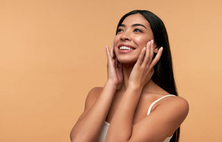 Smiling woman with clear, radiant skin touching her face against a warm background, representing Mirabella Beauty All-In-One Clarifying Serum and healthy glow.