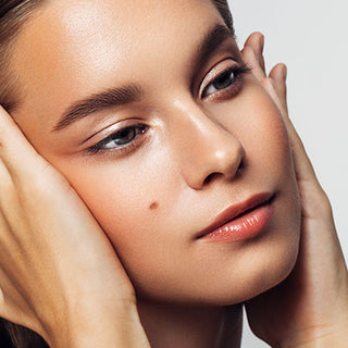 Close-up of Mirabella model on light gray background with hands caressing face & looking upward wearing Invincible For All Anti Aging HD Foundation.
